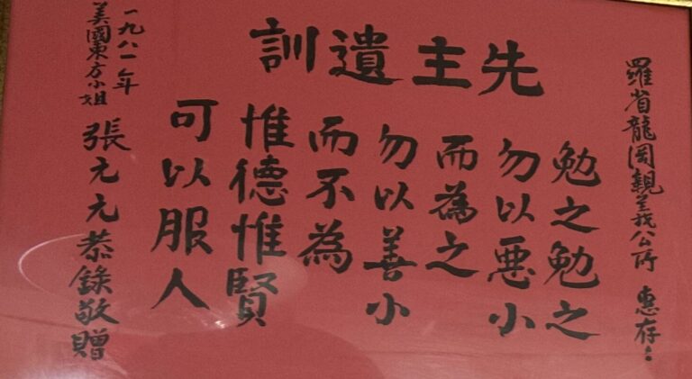 Lung Kong Tin Yee Association motto printed out in a frame on a wall next to two other framed papers and above an exit sign.